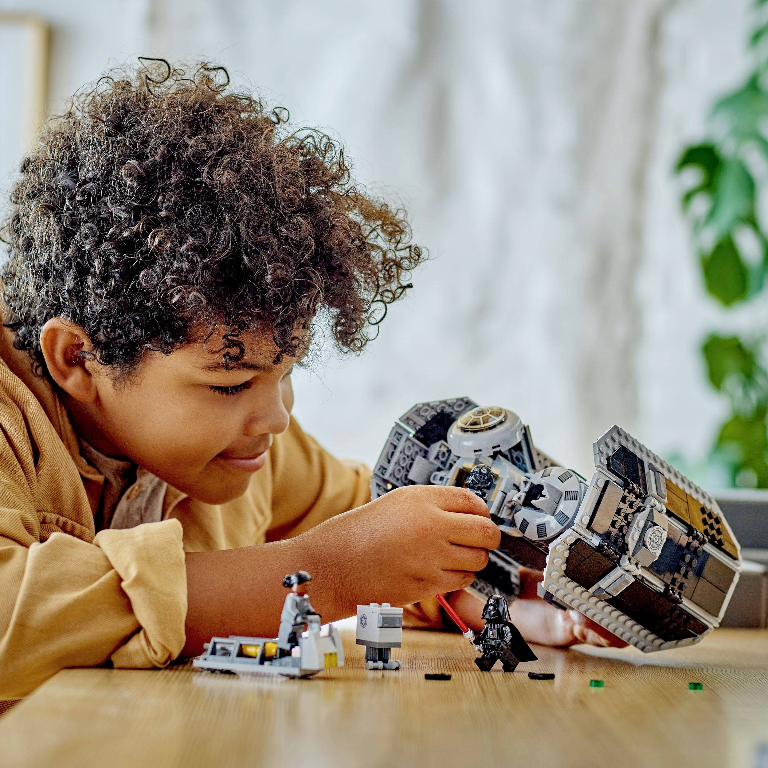 A child is playing on a table with a spaceship and figurine construction kit. They appear focused and creative. Plants are visible in the background.
