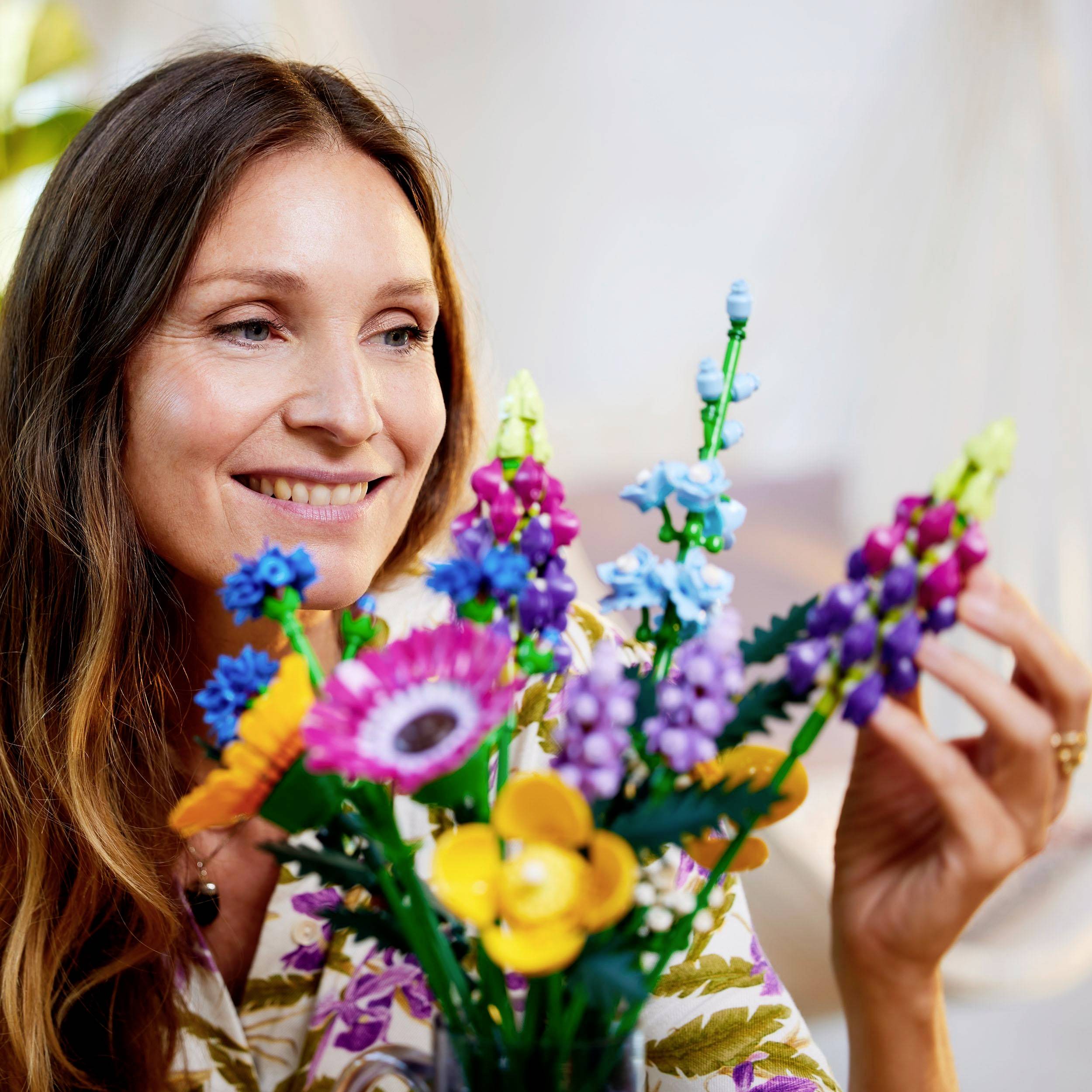A woman smiles as she looks at a colourful bouquet made of LEGO bricks. Blurred plants are visible in the background.