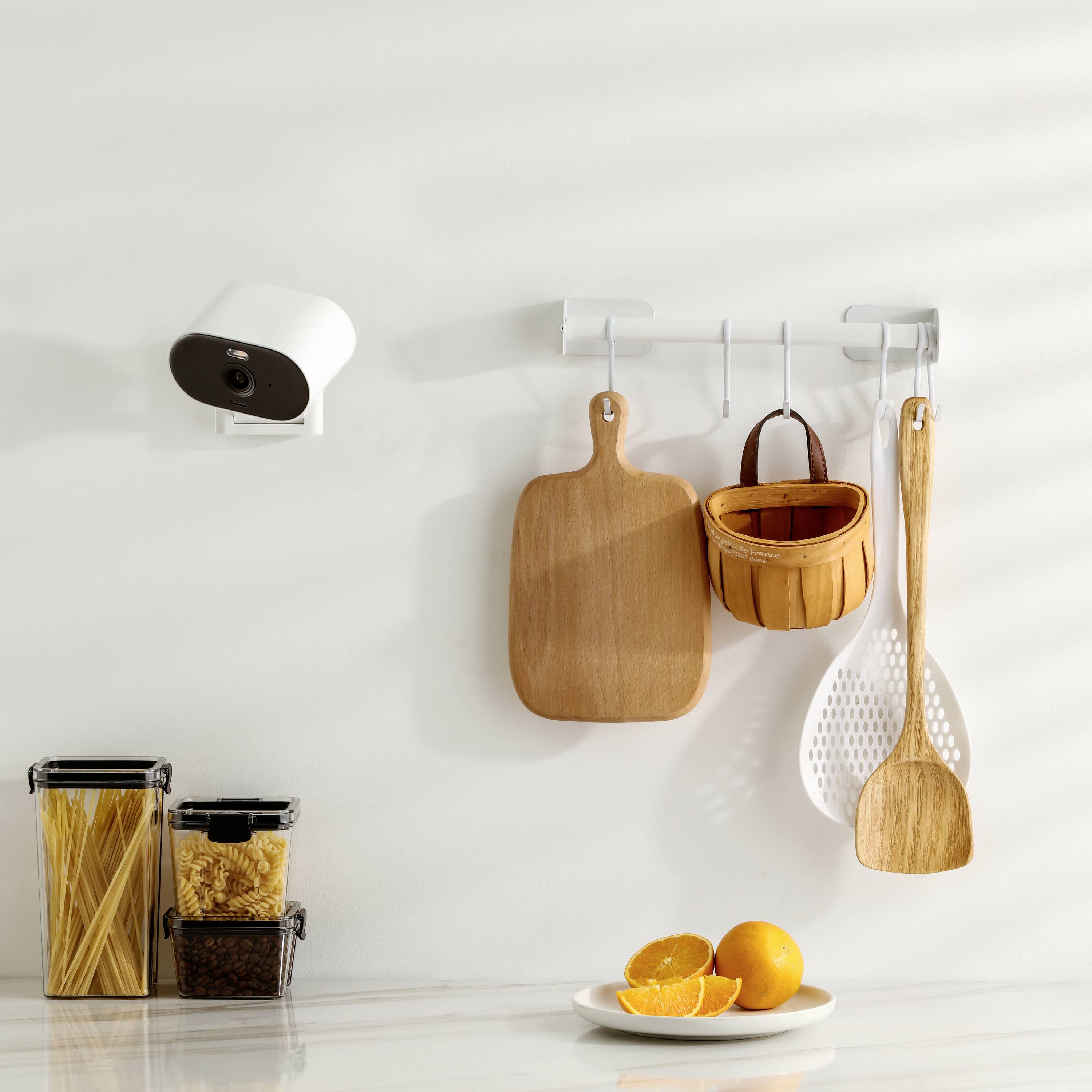 Kitchen with camera mounted on the wall, chopping board, wooden spoon and preserve jars containing pasta and rice on a light worktop.