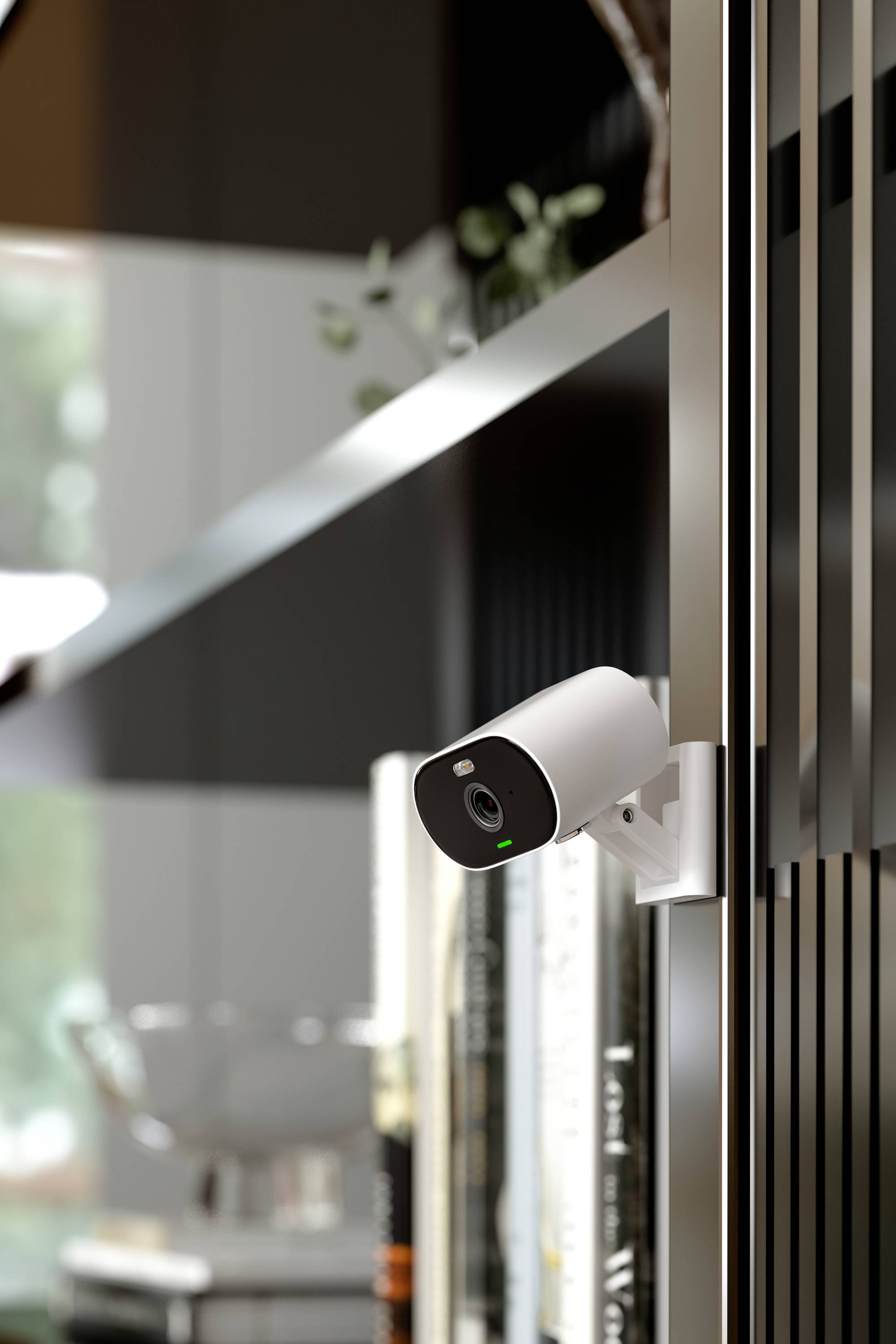A security camera is mounted on a shelf. Books and blurred interior details can be seen in the background.