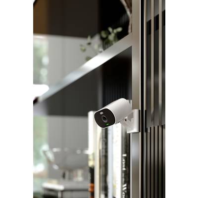 A security camera is mounted on a shelf. Books and blurred interior details can be seen in the background.