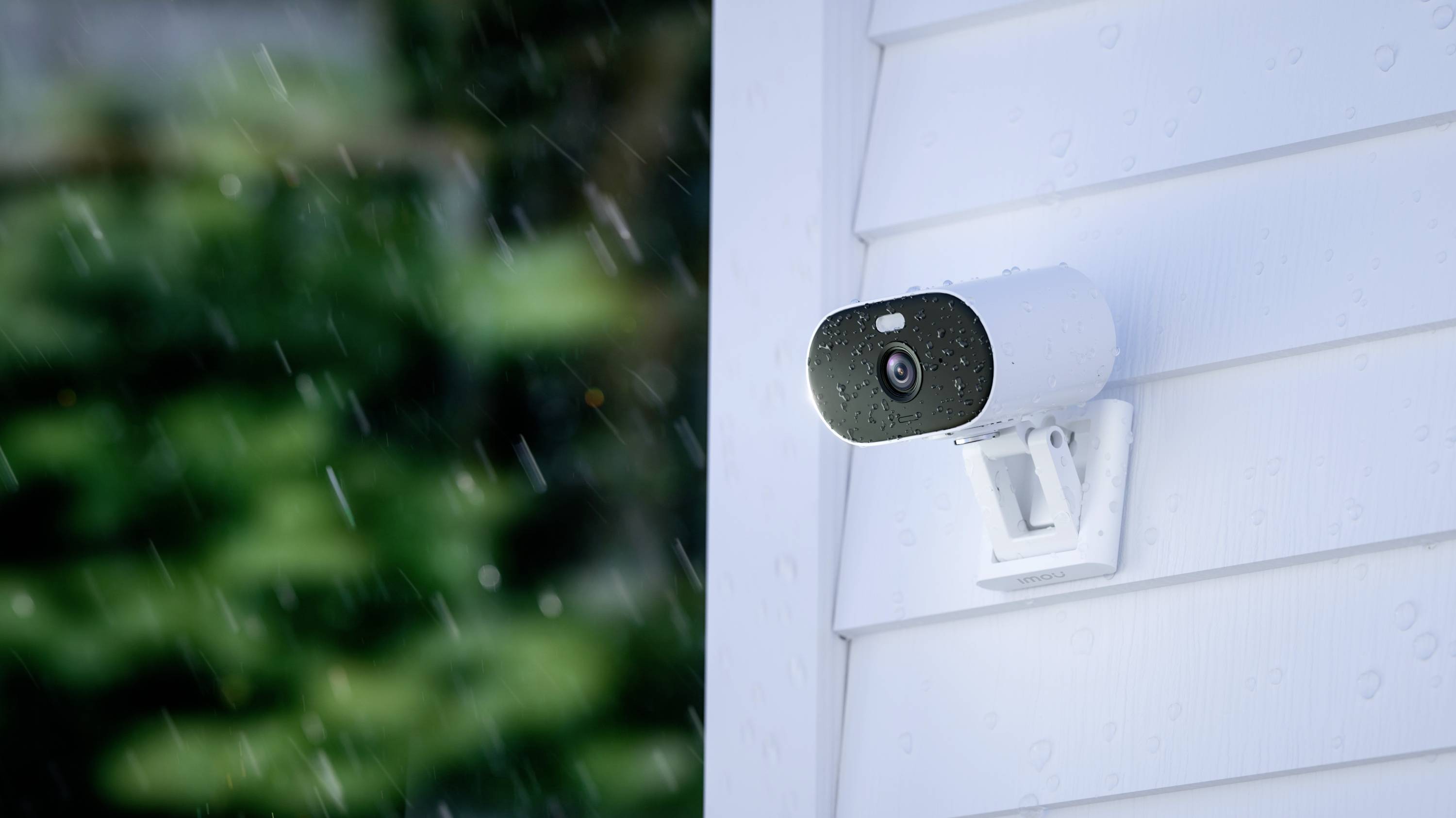 Security camera mounted on a white wall in the rain, focused to the right side. Background shows blurred, green vegetation.