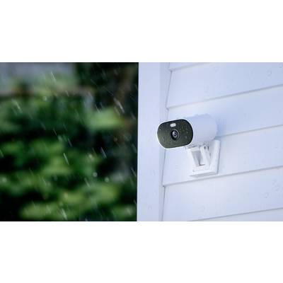 Security camera mounted on a white wall in the rain, focused to the right side. Background shows blurred, green vegetation.