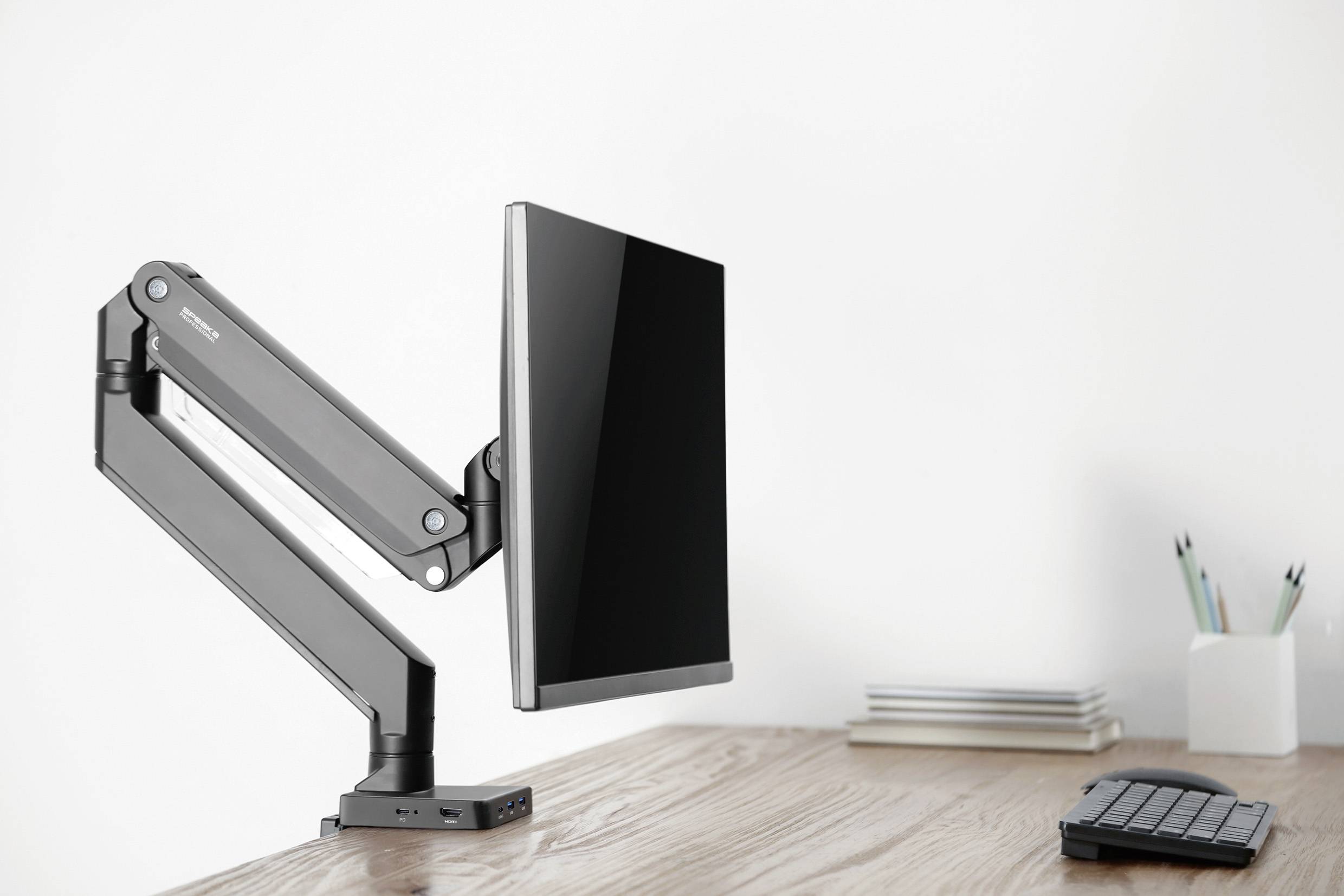 Monitor arm with a mounted screen on a wooden desk, beside a keyboard and writing tools; minimalist office setup.