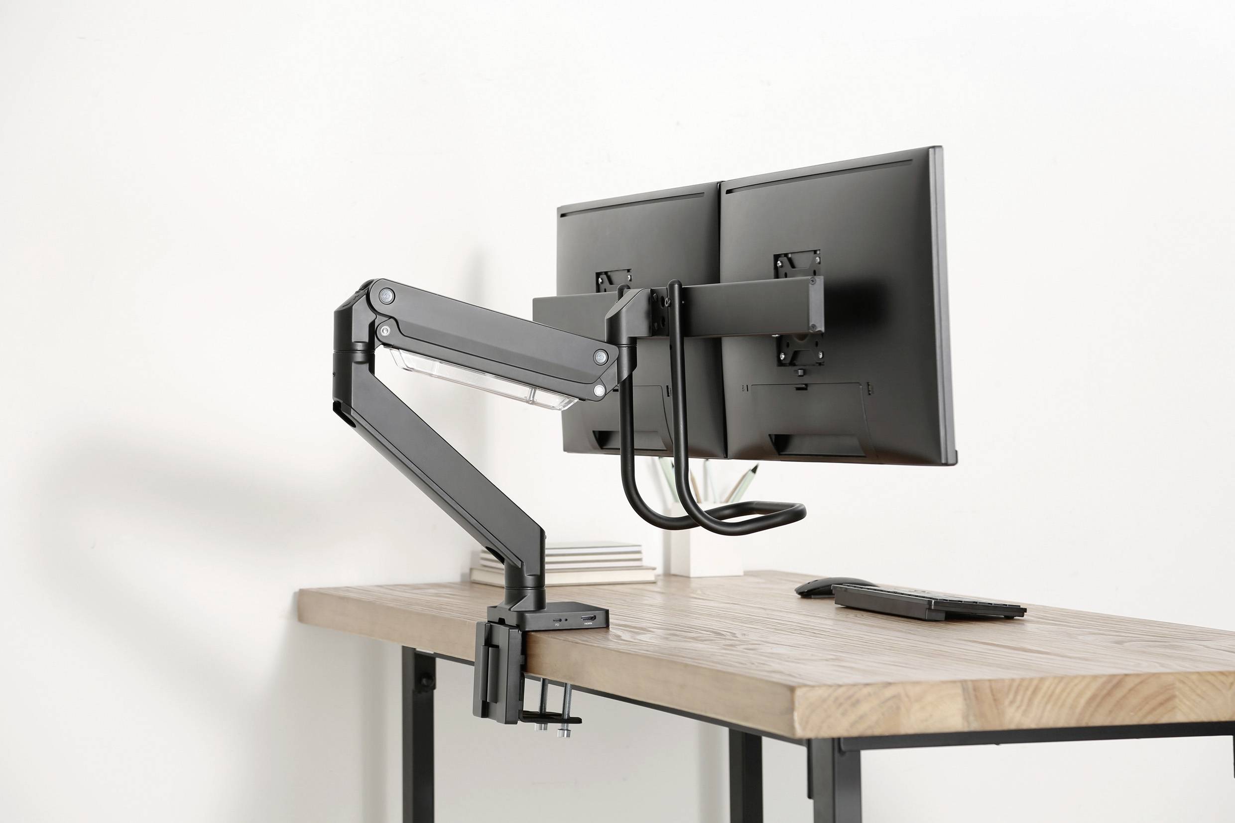 A desk with a dual monitor arm mount, on which two monitors are attached. The desk is made of wood and has a modern design.