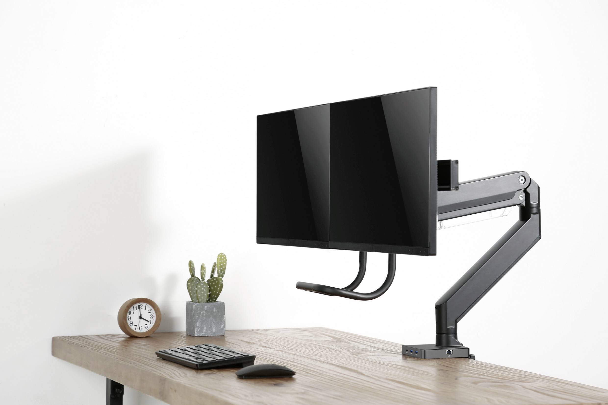 A computer monitor with an arm mount on a desk next to a keyboard, a cactus, and an alarm clock.