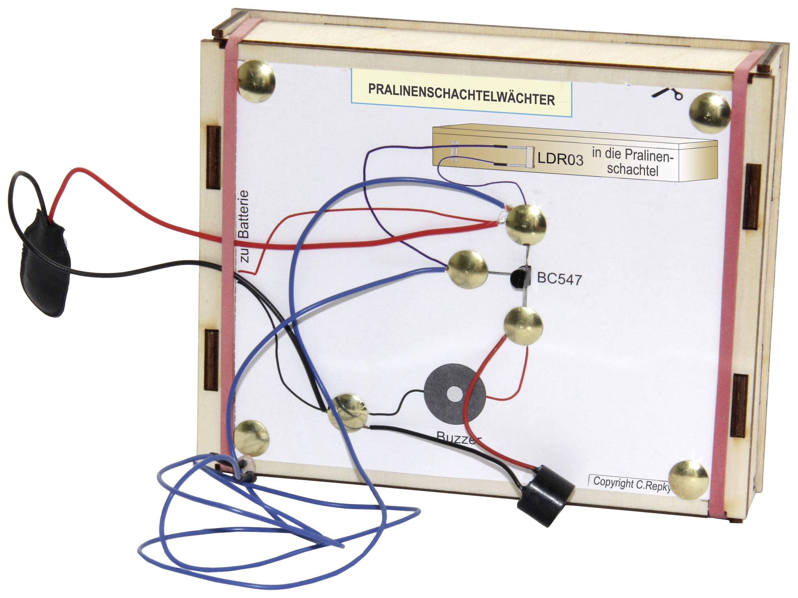 Electronic circuit on a wooden board for monitoring a chocolate box; wires, battery connection, and a sensor are visible.