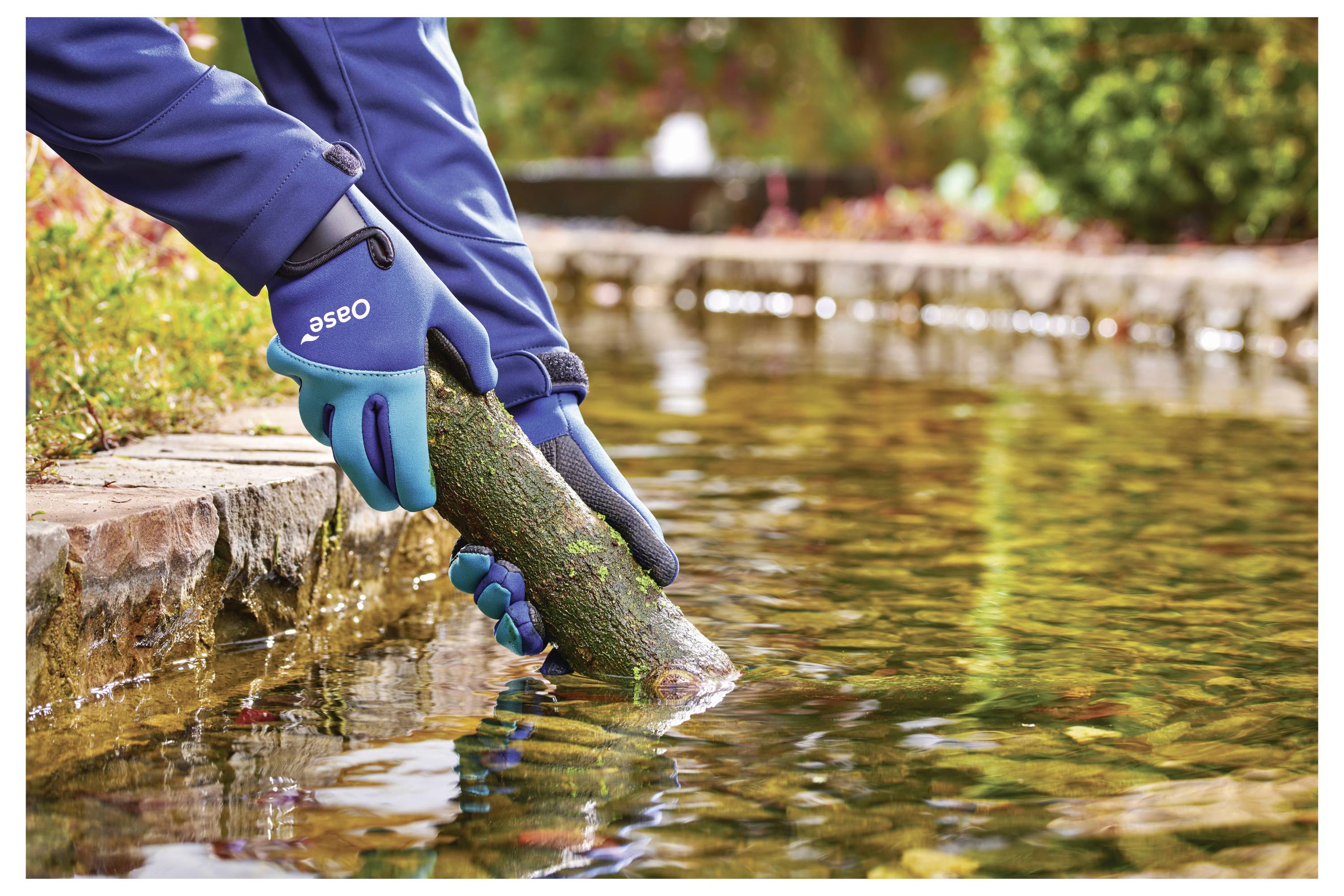 Hands wearing gloves placing a mossy log into a pond, surrounded by autumn leaves and a stone edge, suggesting a garden scene.