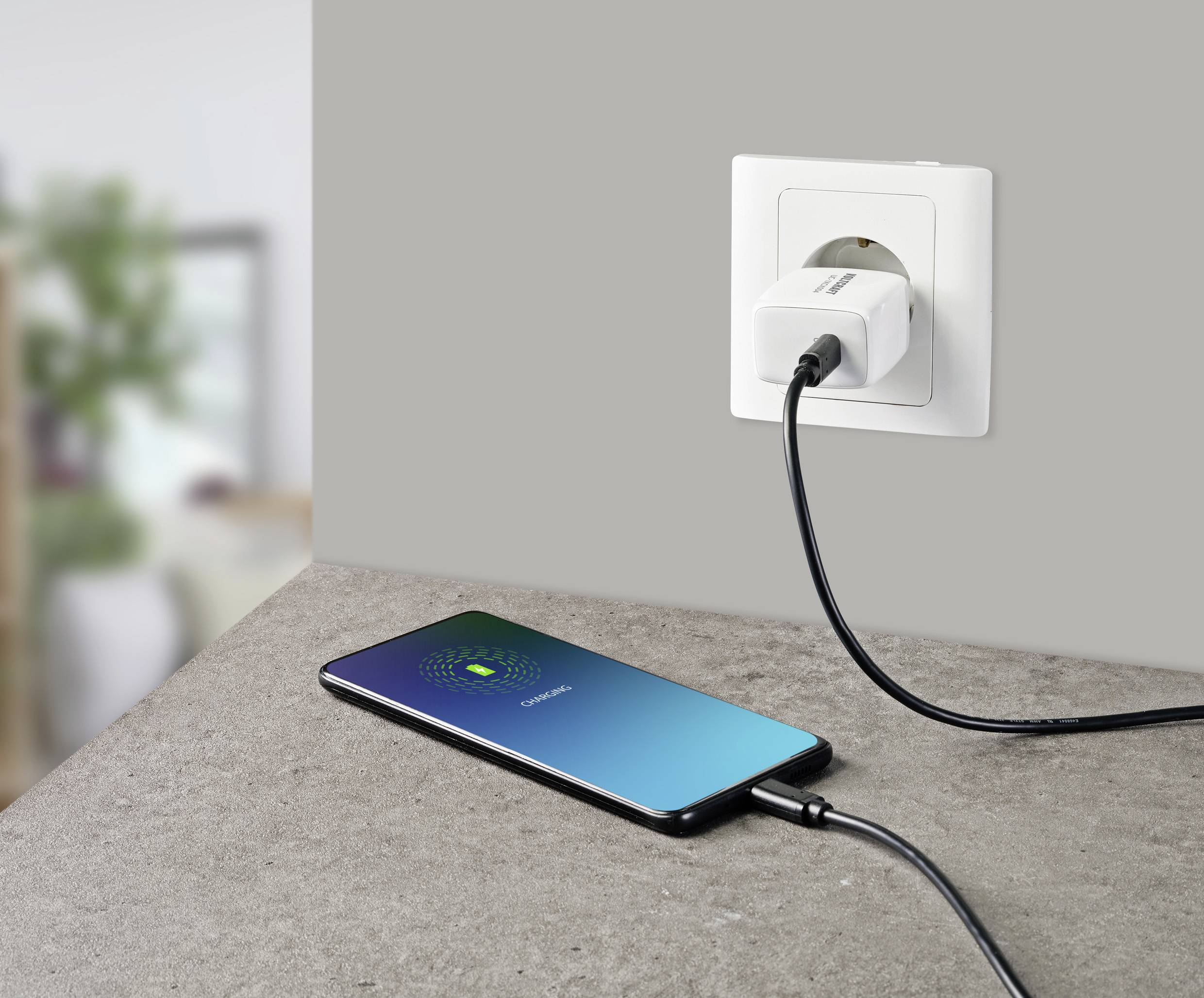 A mobile phone is lying on a table and is connected to a power socket with a cable. The screen displays 'Charging'.