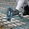 A person is securing steel reinforcements on a construction site with a blue cordless device. They are wearing work gloves and white workwear.