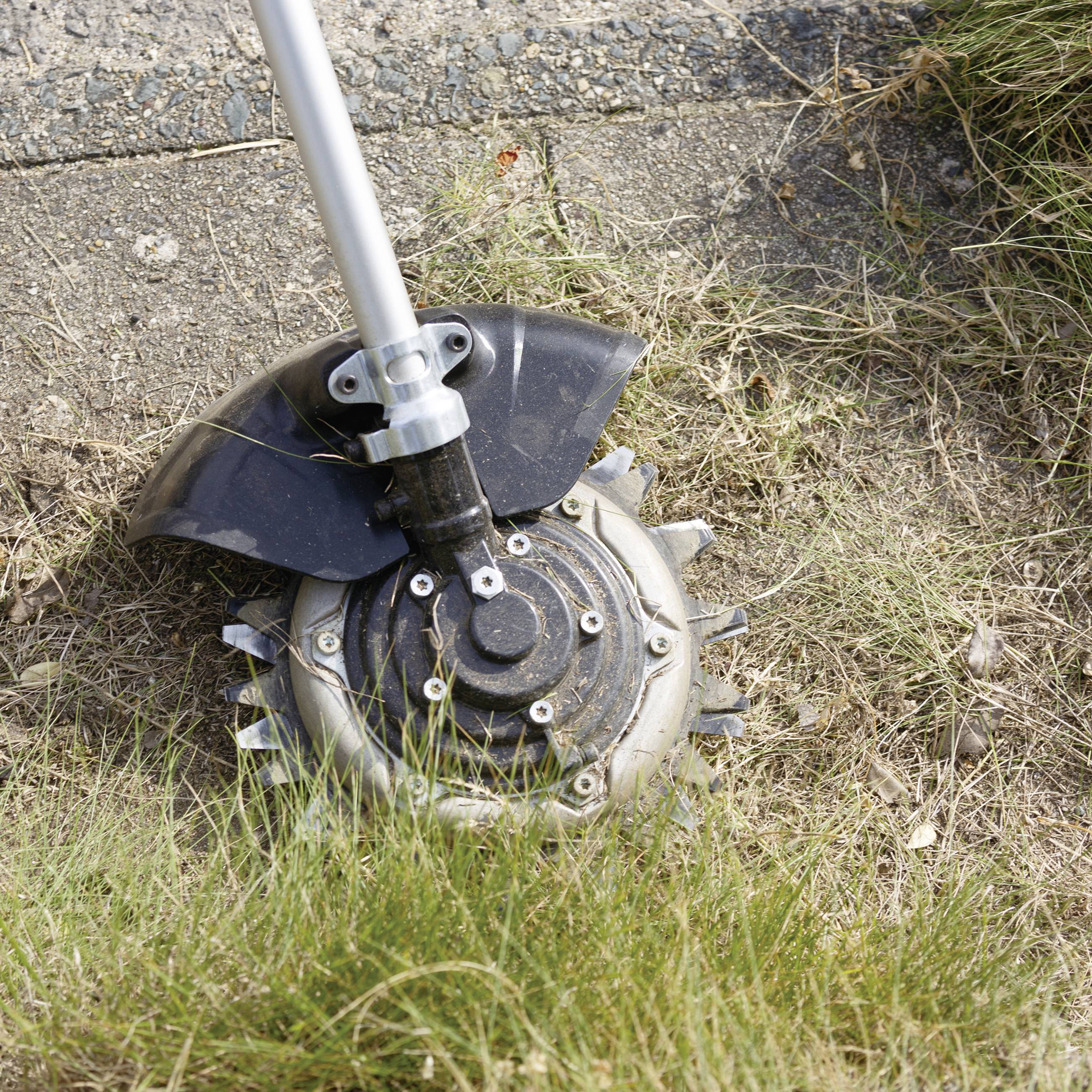 A lawn trimmer head lies on dry grass and earth beside a pavement, ready for use.