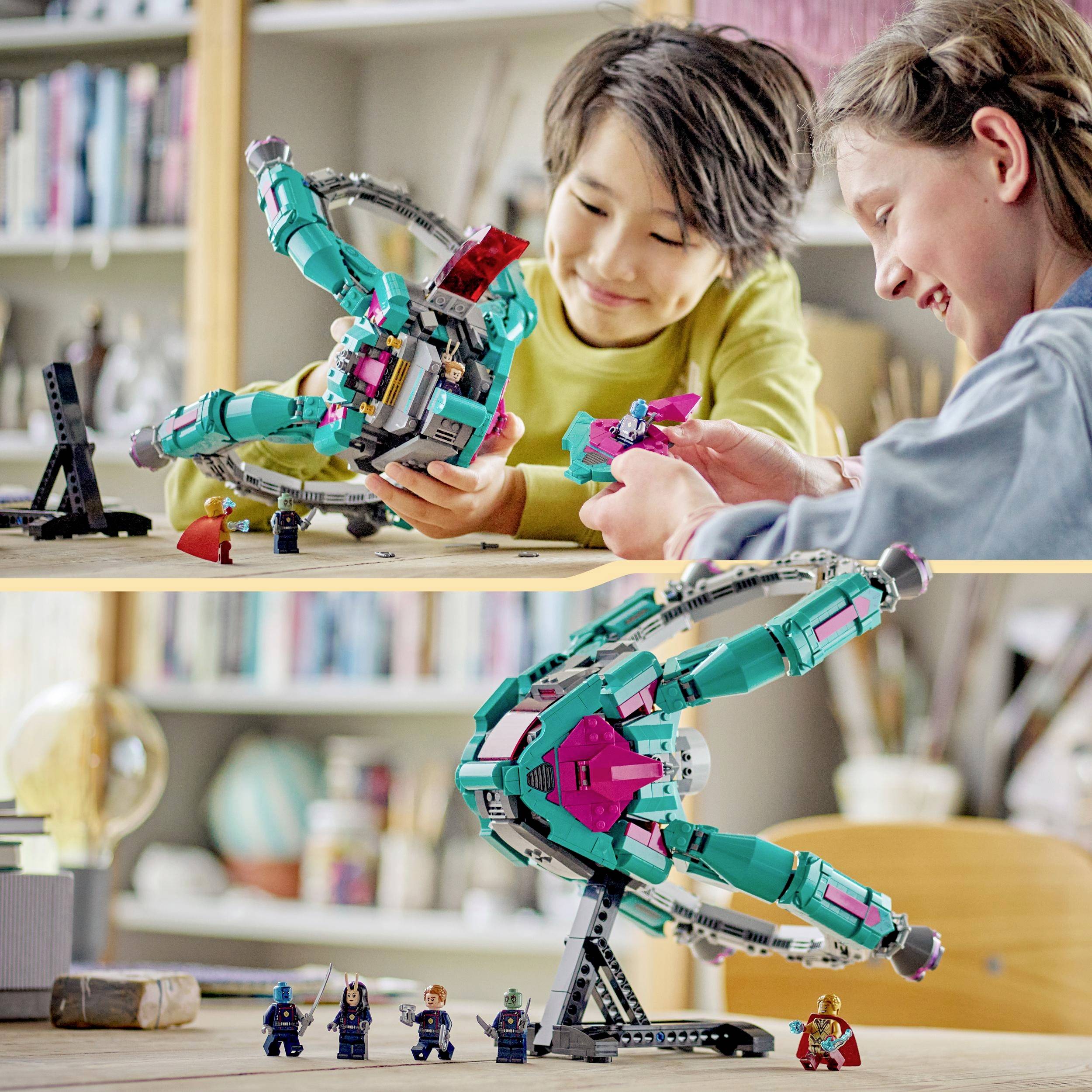 Two children are happily playing with a colourful spaceship model and small figures on a table, surrounded by books and shelves.