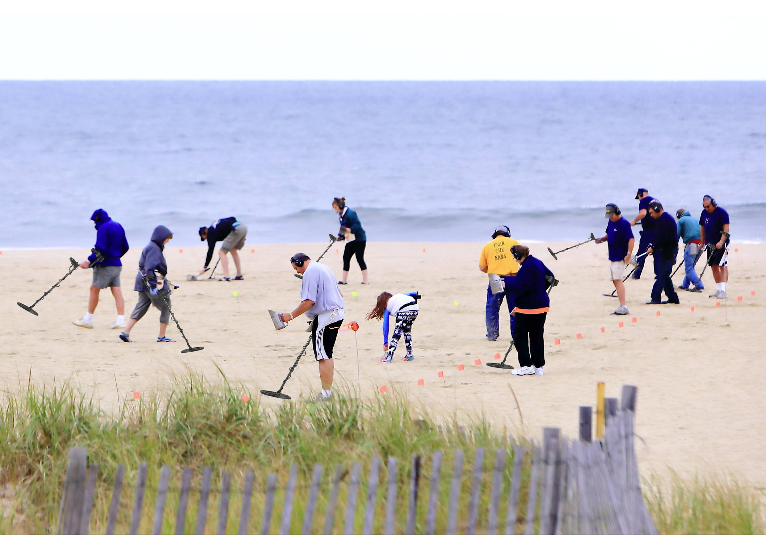 A group of people searching for treasures with metal detectors on the beach. Sand, sea, and clouds in the background.