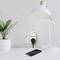 A mobile phone is connected to a power socket with a charging cable. Next to it stands a table lamp and a plant.