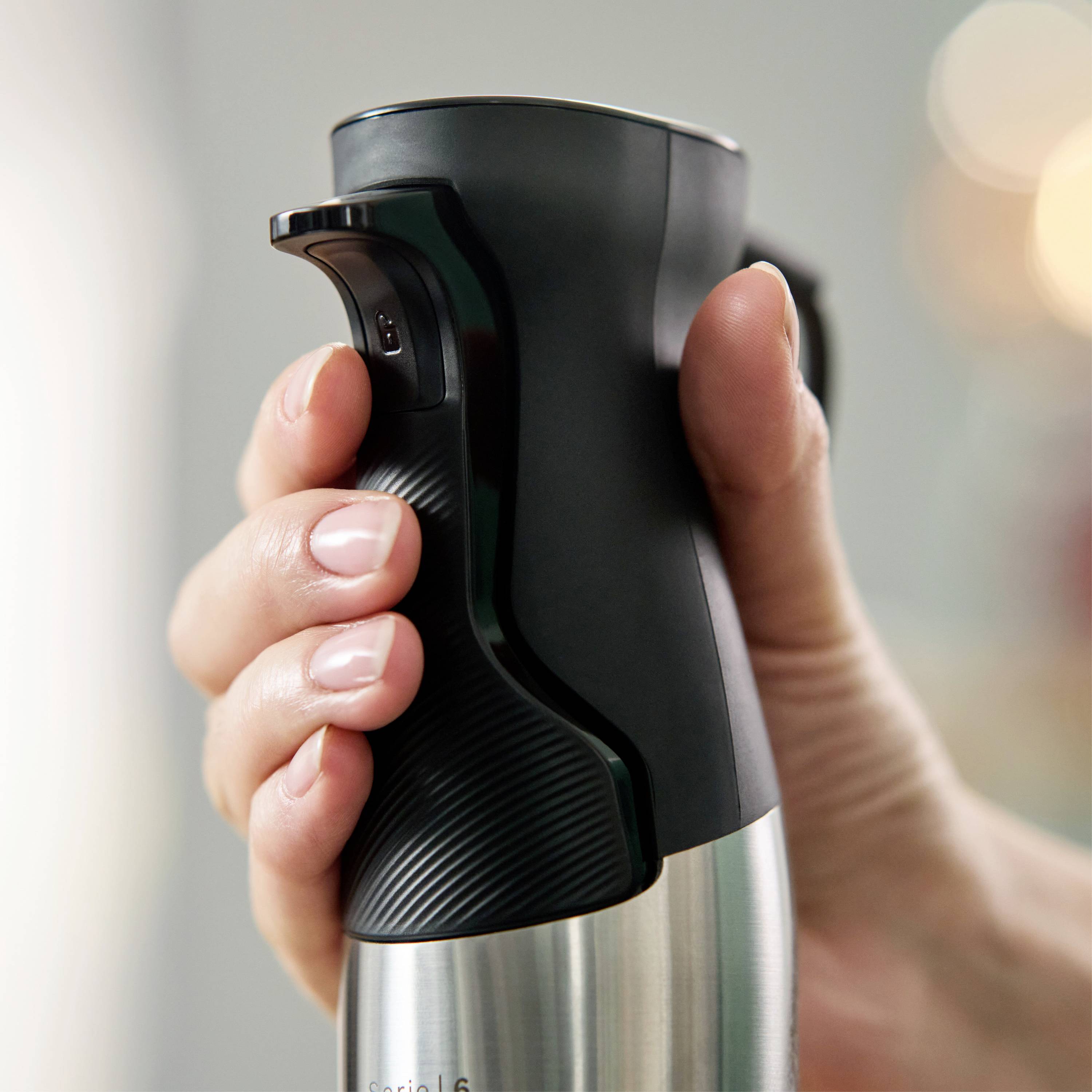 A hand is holding a large stainless steel thermos flask. The focus is on the black handle and the lever for pouring.