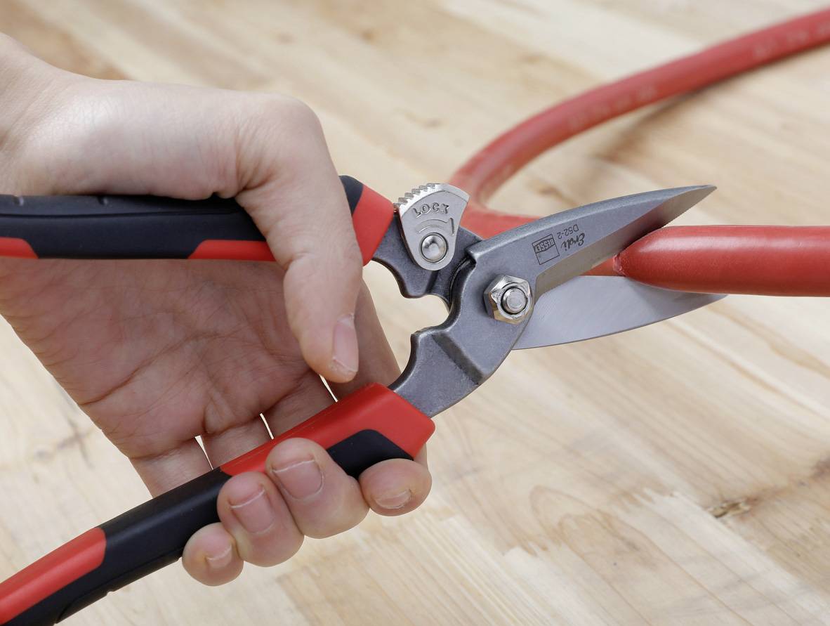 A hand holds secateurs cutting through a red cable. The blade is sharp and firmly closed around the cable.