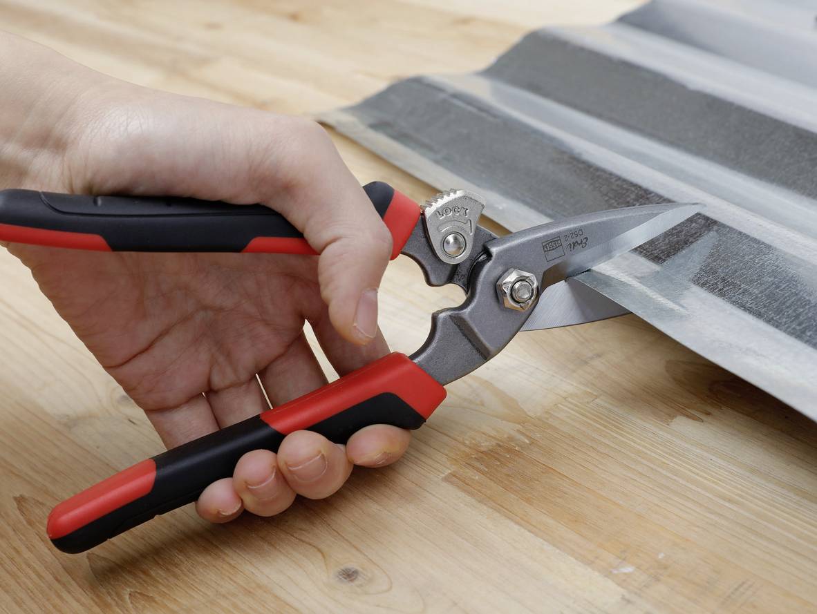 A person is cutting corrugated metal sheeting with tin snips on a wooden table.