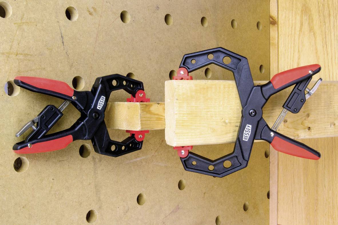 Two red and black clamps are holding a piece of wood securely in place on a perforated board to stabilise it.