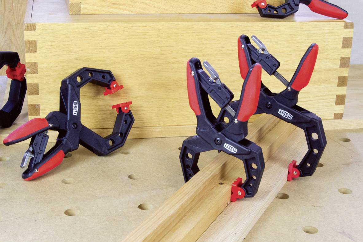 Two plastic clamps with red handles are holding wooden pieces on a workbench, against a background of wooden boxes.