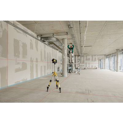 A construction worker stands on scaffolding and is installing pipes on the ceiling in an unfinished room. A laser device is projecting red lines onto the floor.