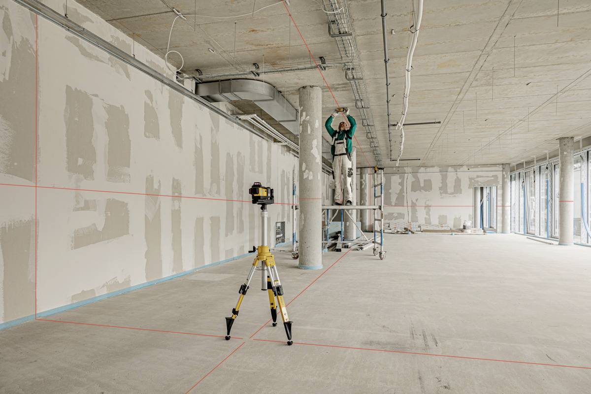 A construction worker is standing on a ladder in a partially completed room, measuring the ceiling height, while a laser level projects lines.