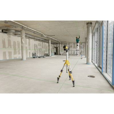 Large empty space in a building under construction, a worker is installing lighting on the ceiling, while a laser level stands on the floor.