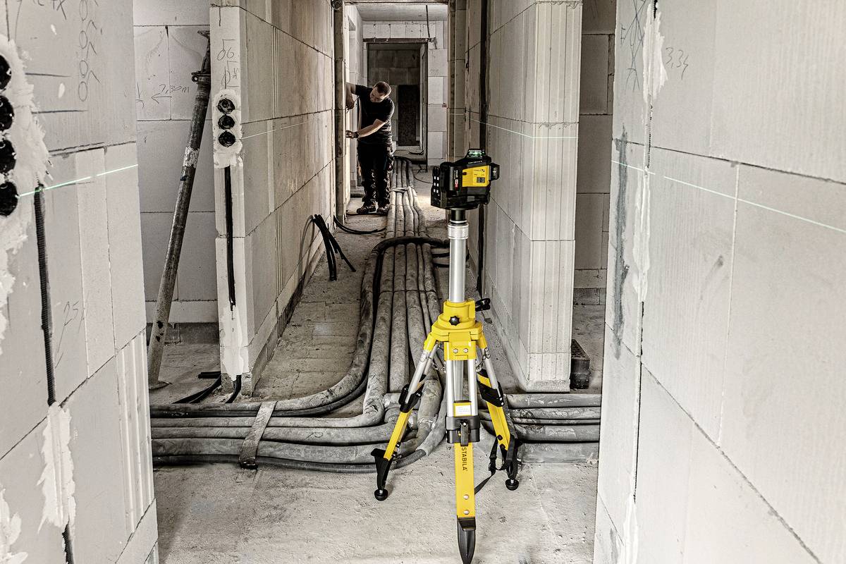 A construction site inside a building with a surveying instrument mounted on a tripod in the foreground. A worker can be seen in the background.