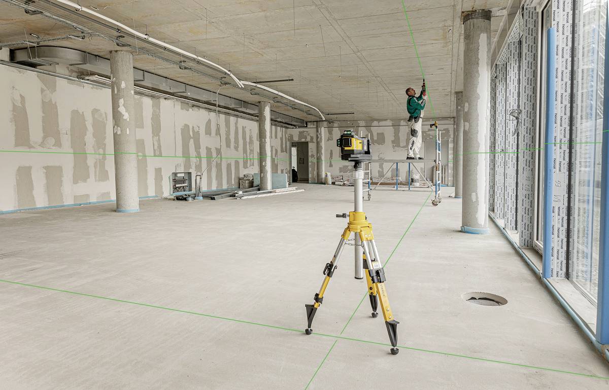 A construction worker in an empty room is aligning a device on the ceiling. A laser levelling tool is positioned on a tripod on the floor.