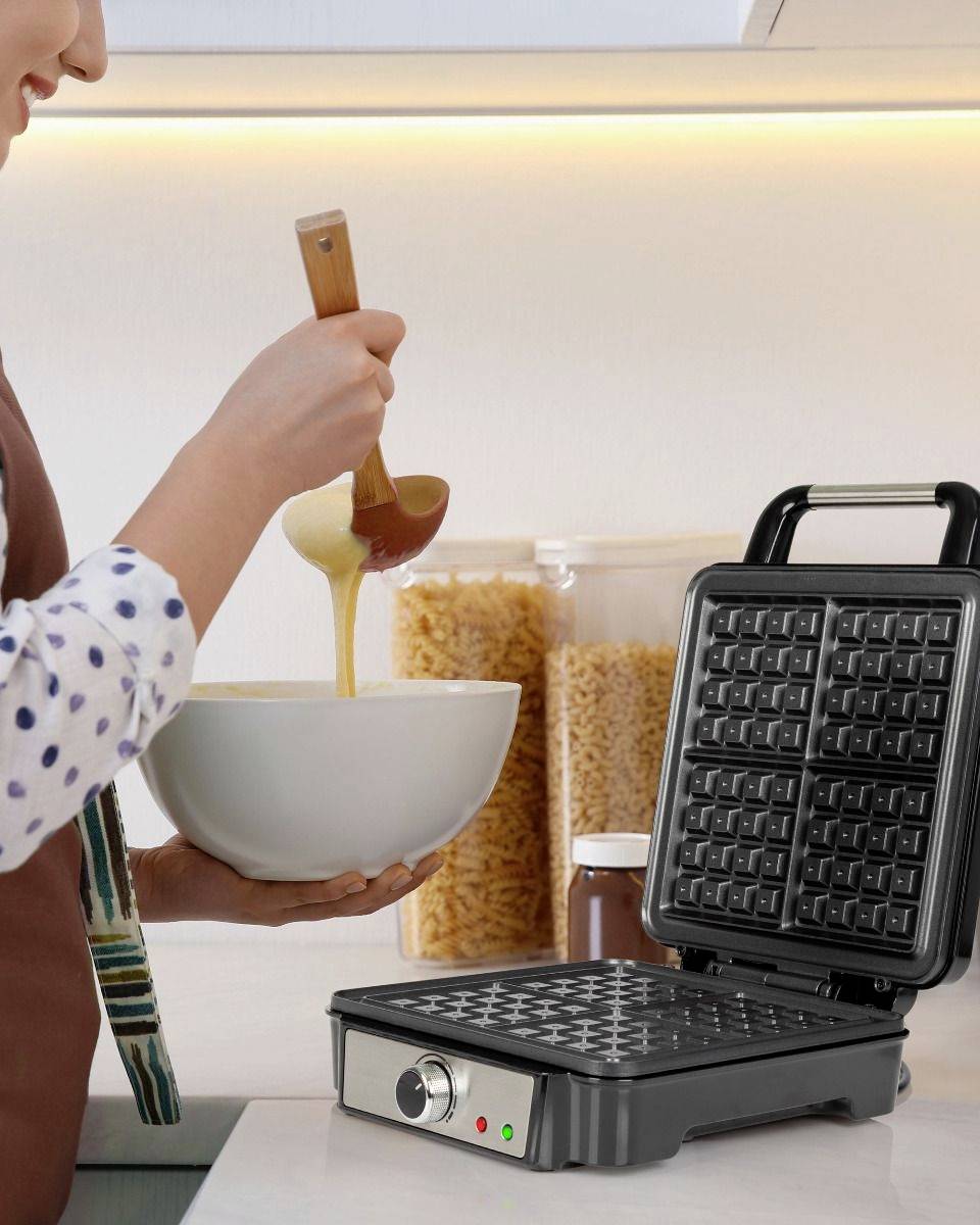 A person is pouring batter into a waffle iron in a kitchen. Storage jars are standing nearby.