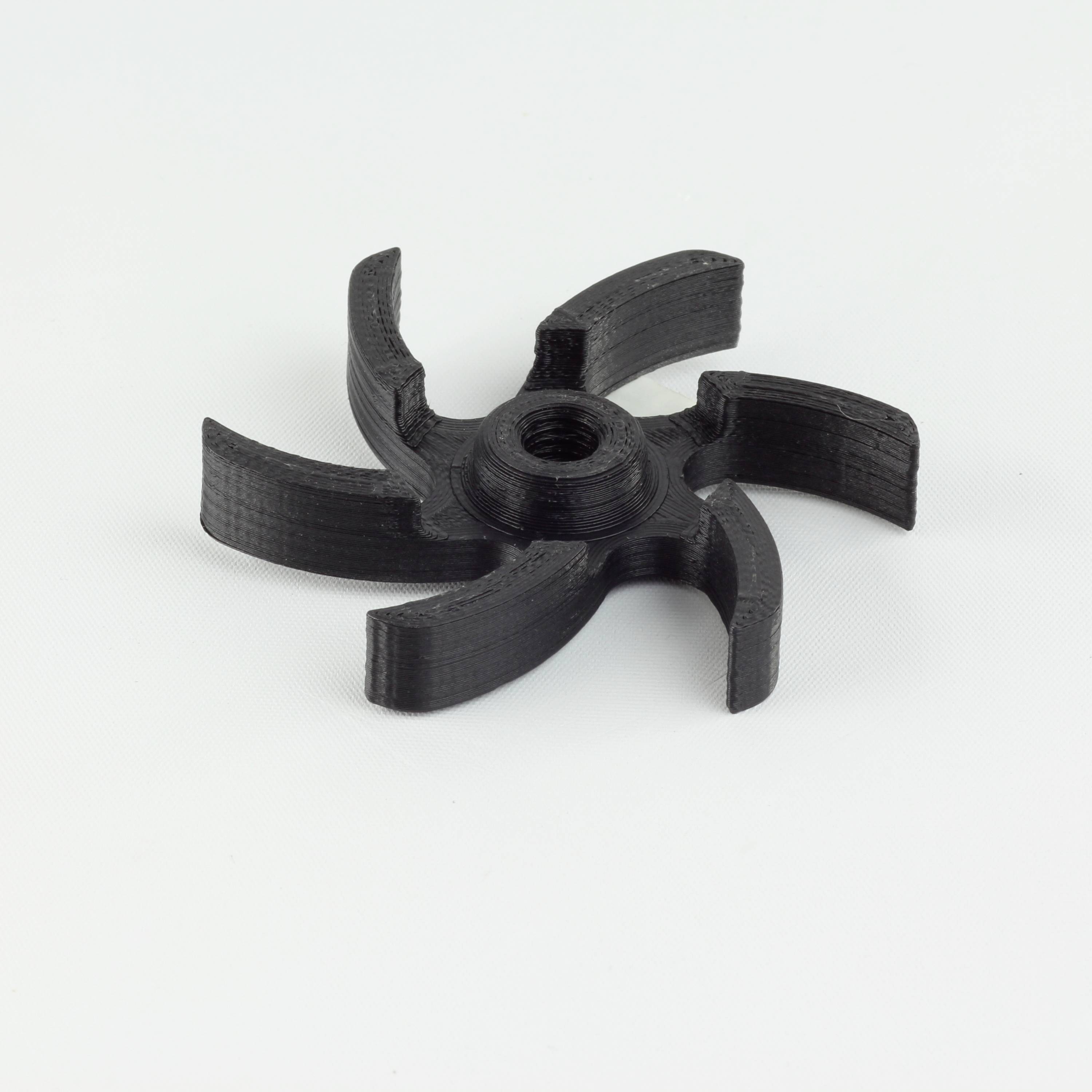 A black plastic part with six curved arms, resembling a rotor or propeller, is lying on a white surface.