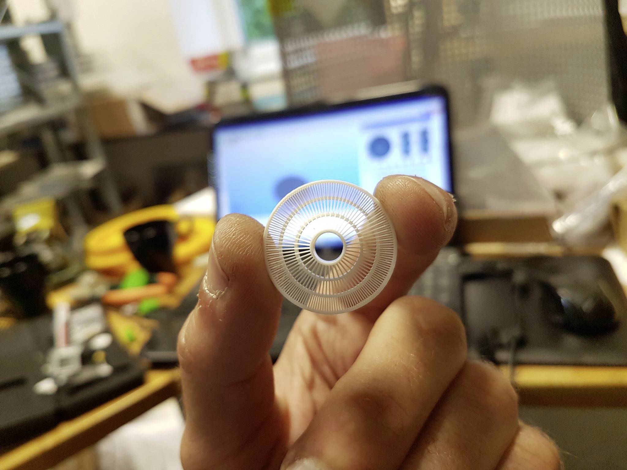 'Close-up of a hand holding a small, round metal filter in front of a blurred laptop screen. Office in the background.'