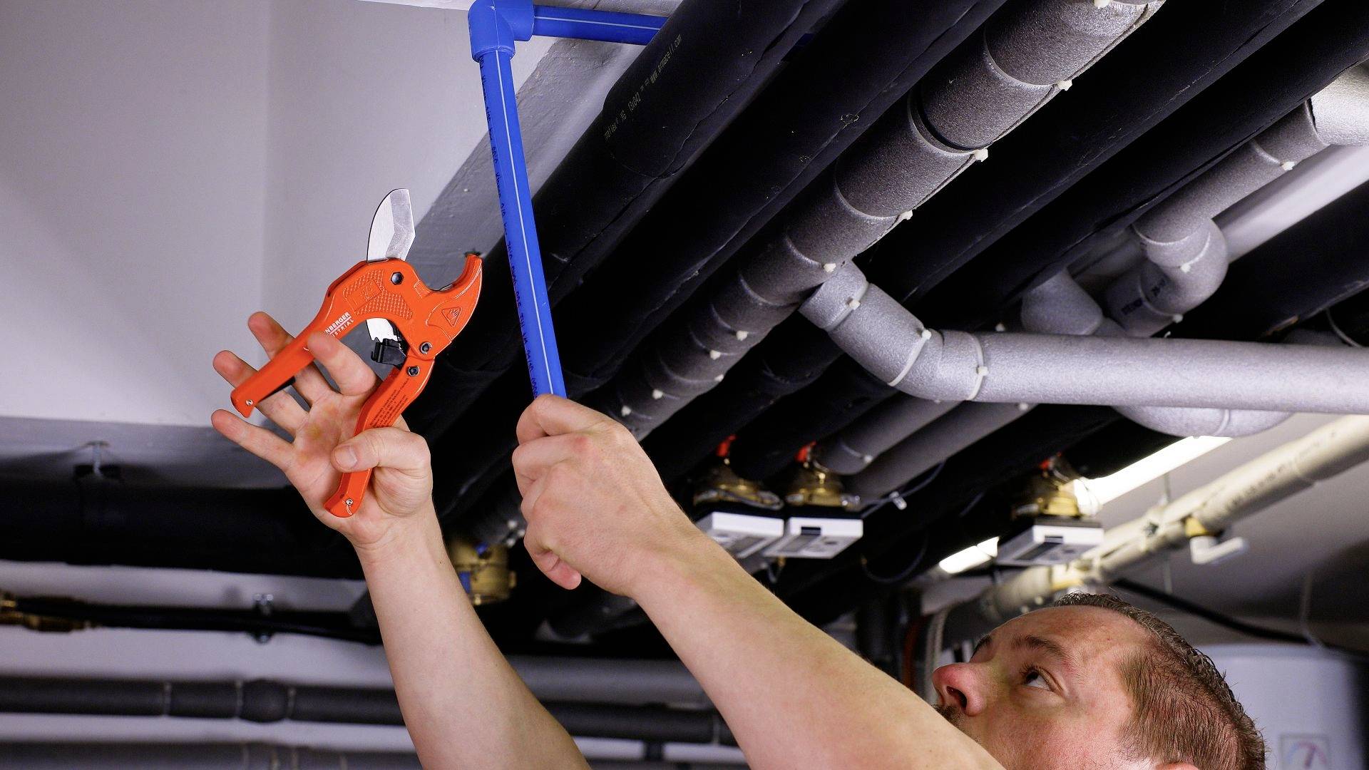 A person is repairing pipes on a ceiling using a red pipe wrench. The background shows several grey and black pipes.