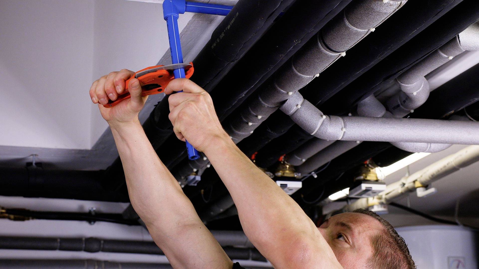 A tradesman is repairing pipes on the ceiling using pliers. The scene demonstrates careful manual work and technical precision.