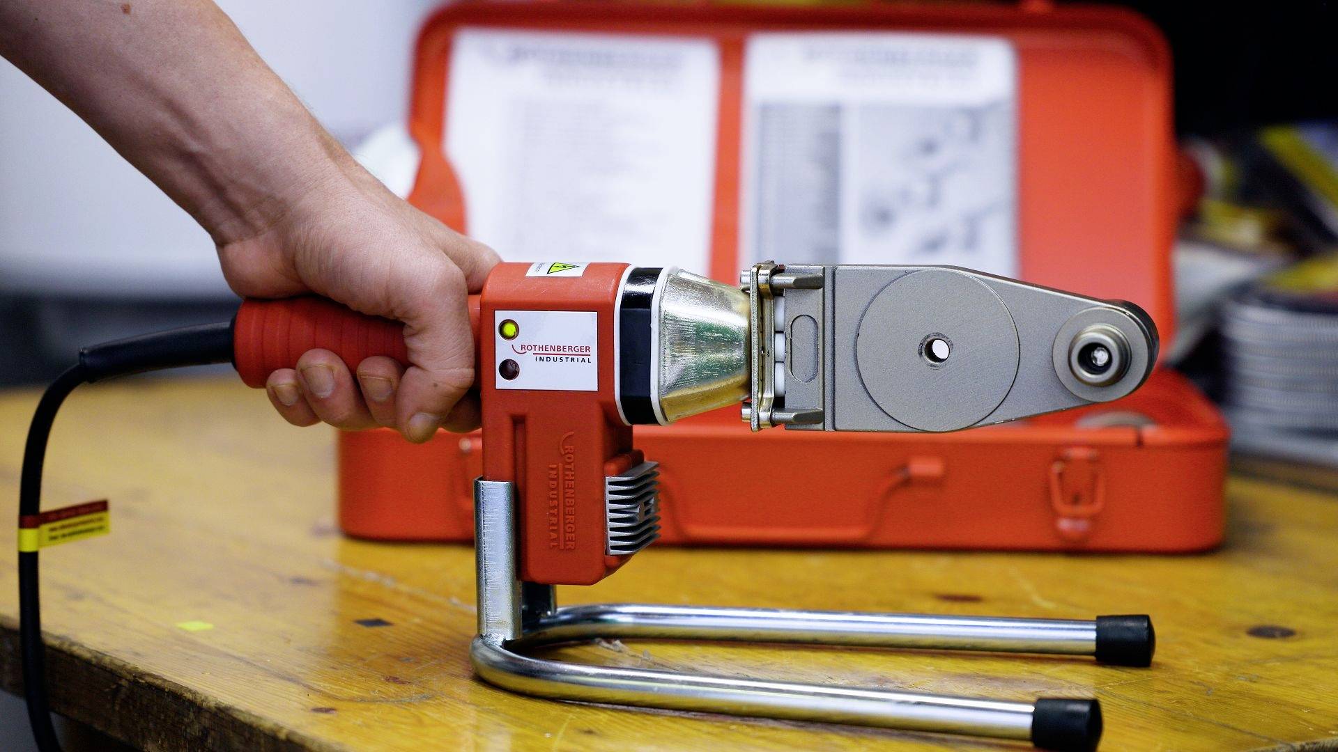 A person is holding a red welding device for plastic pipes. In the background, an open red case with an instruction manual is visible.