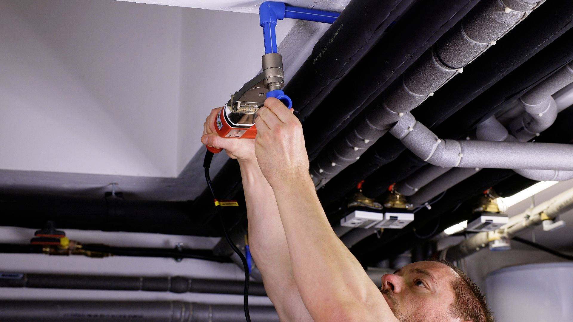 A man is installing or repairing pipes on a ceiling. He is using a tool to connect parts. The pipes are black and blue.