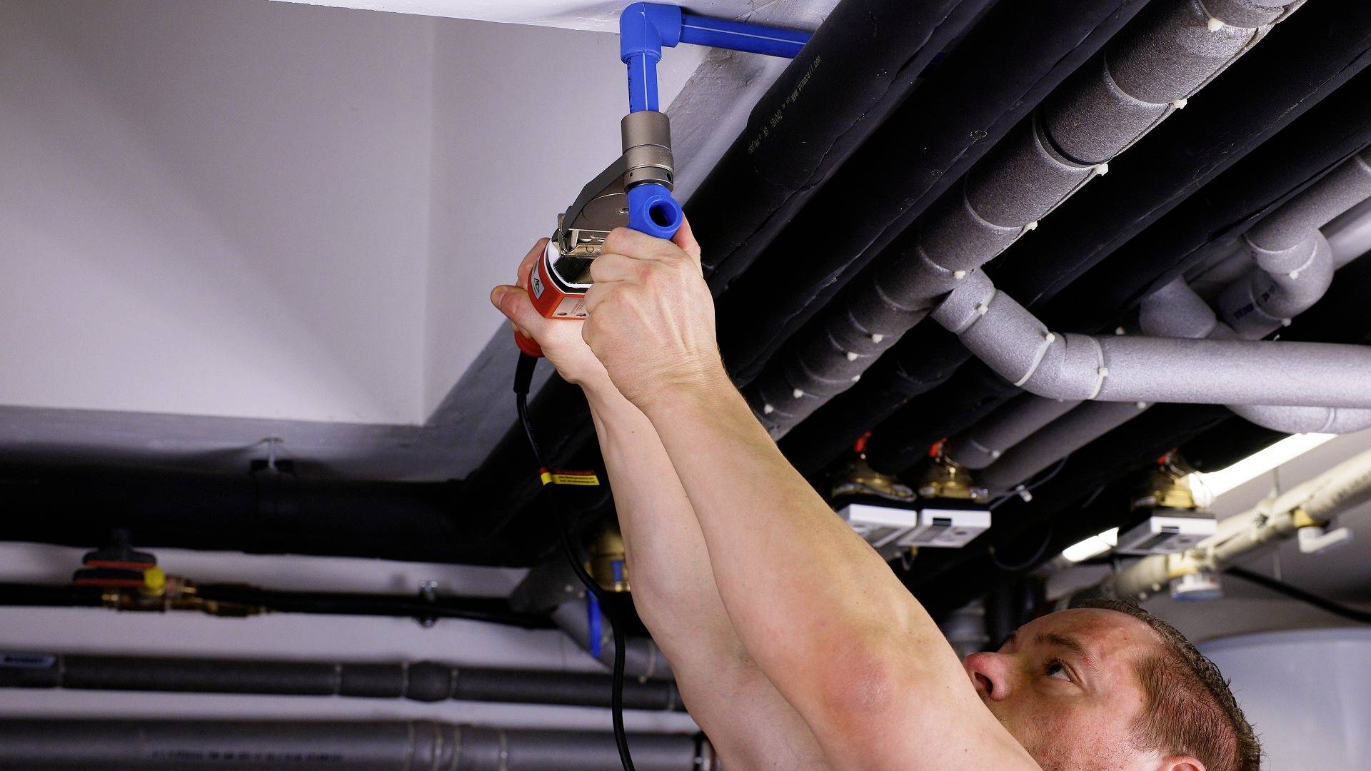 A man is repairing a pipe system beneath a ceiling. He is using tools to work on a blue pipe. Additional pipes are visible in the background.