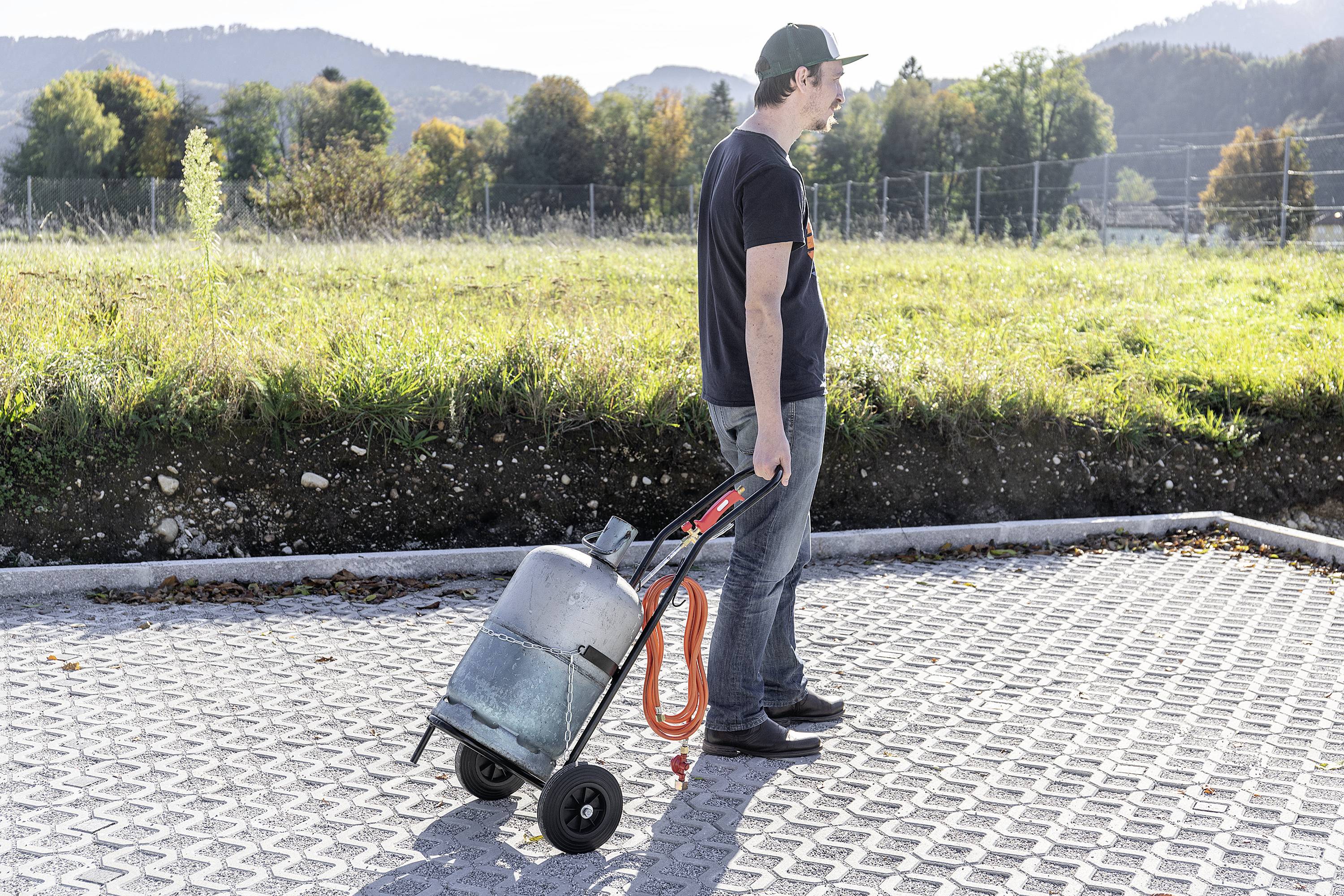 A man is pulling a gas cylinder on a small handcart across a paved car park. Trees and hills can be seen in the background.