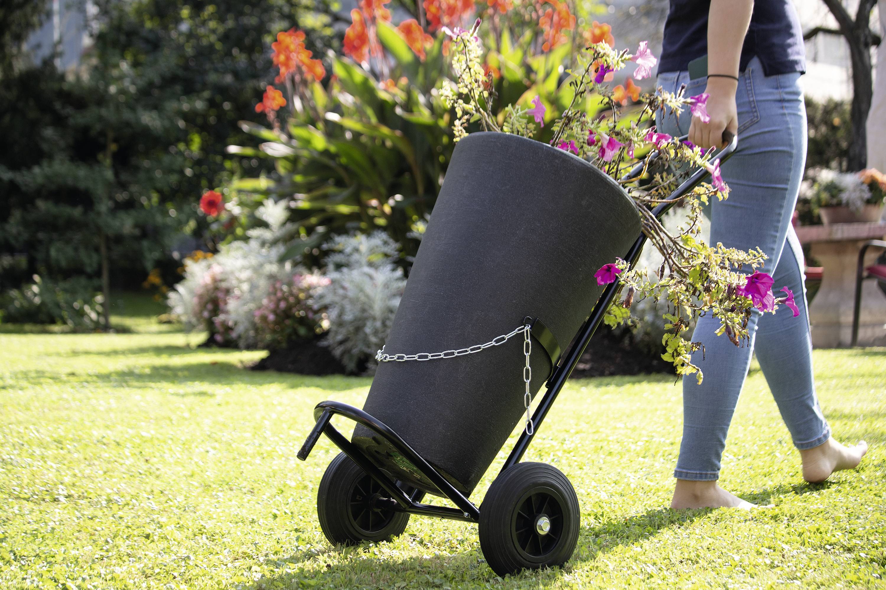 A person is transporting a large basket of flowers on a handcart across a green lawn in the garden.