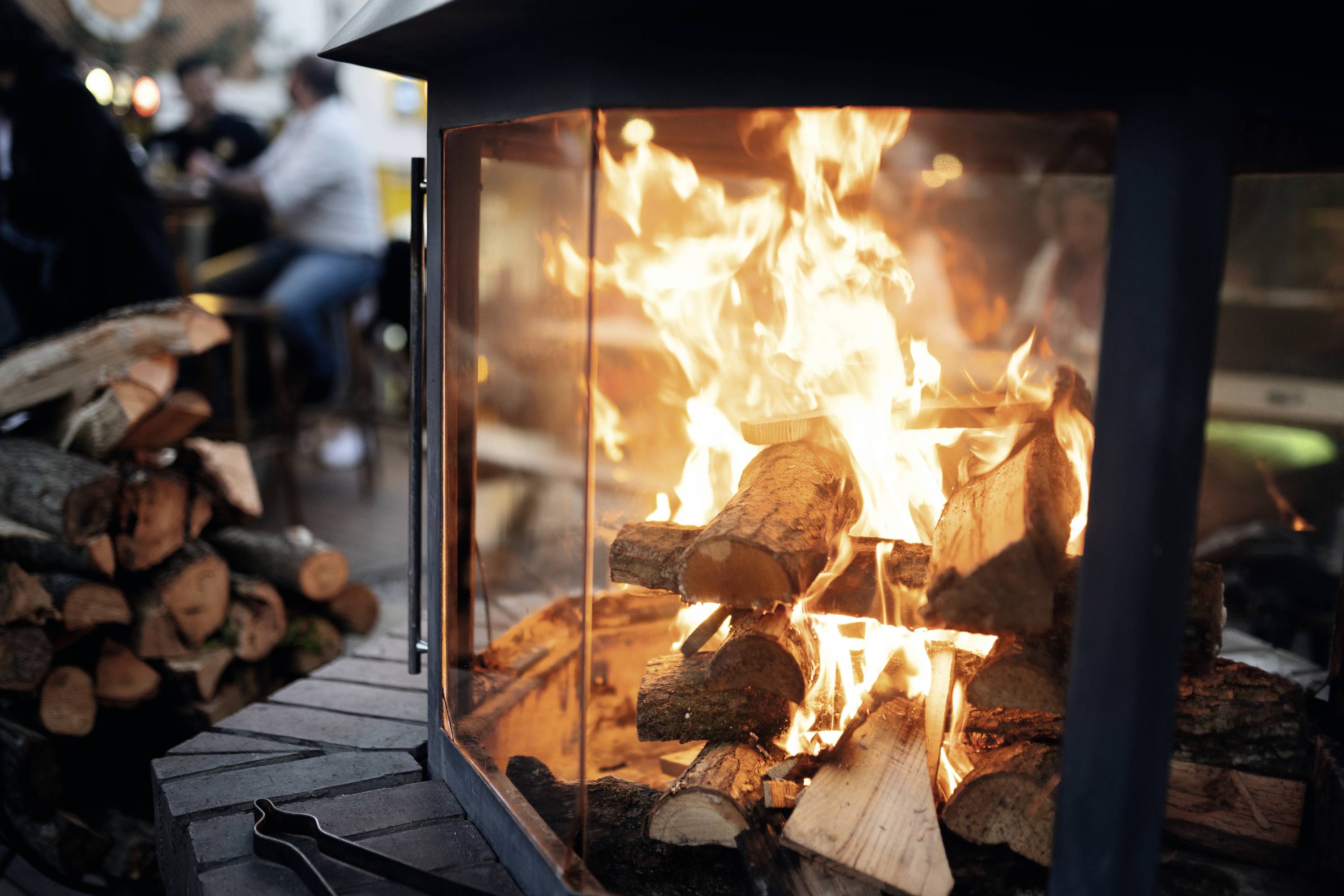 Logs are burning in an open fireplace. People in the background suggest a cosy outdoor area.