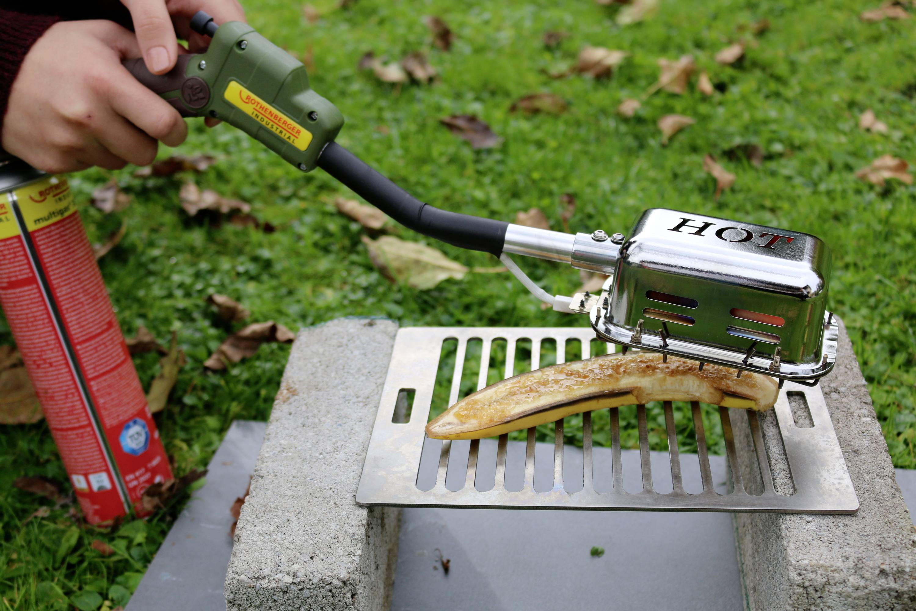 A person is heating a yellow object on a barbecue grill using a blowtorch. Grass and leaves are visible in the background.