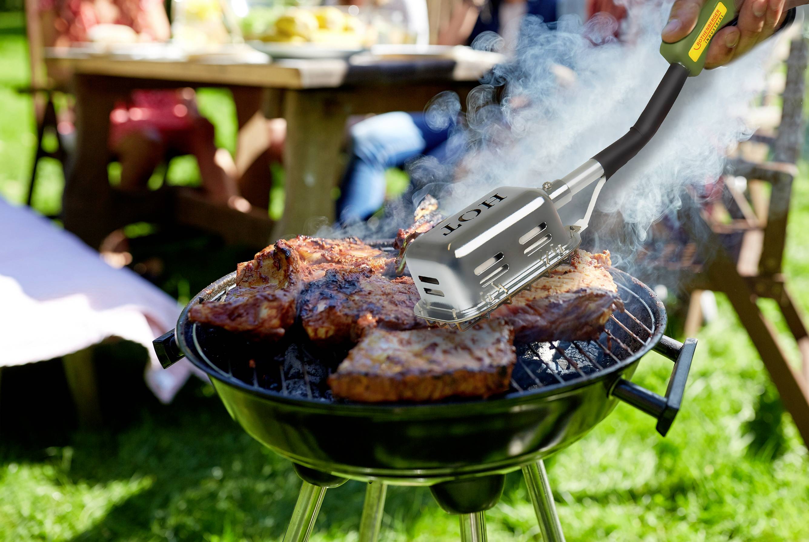 A barbecue with large pieces of meat sizzling. Smoke rises. In the background, a group of people are seated at an outdoor table.