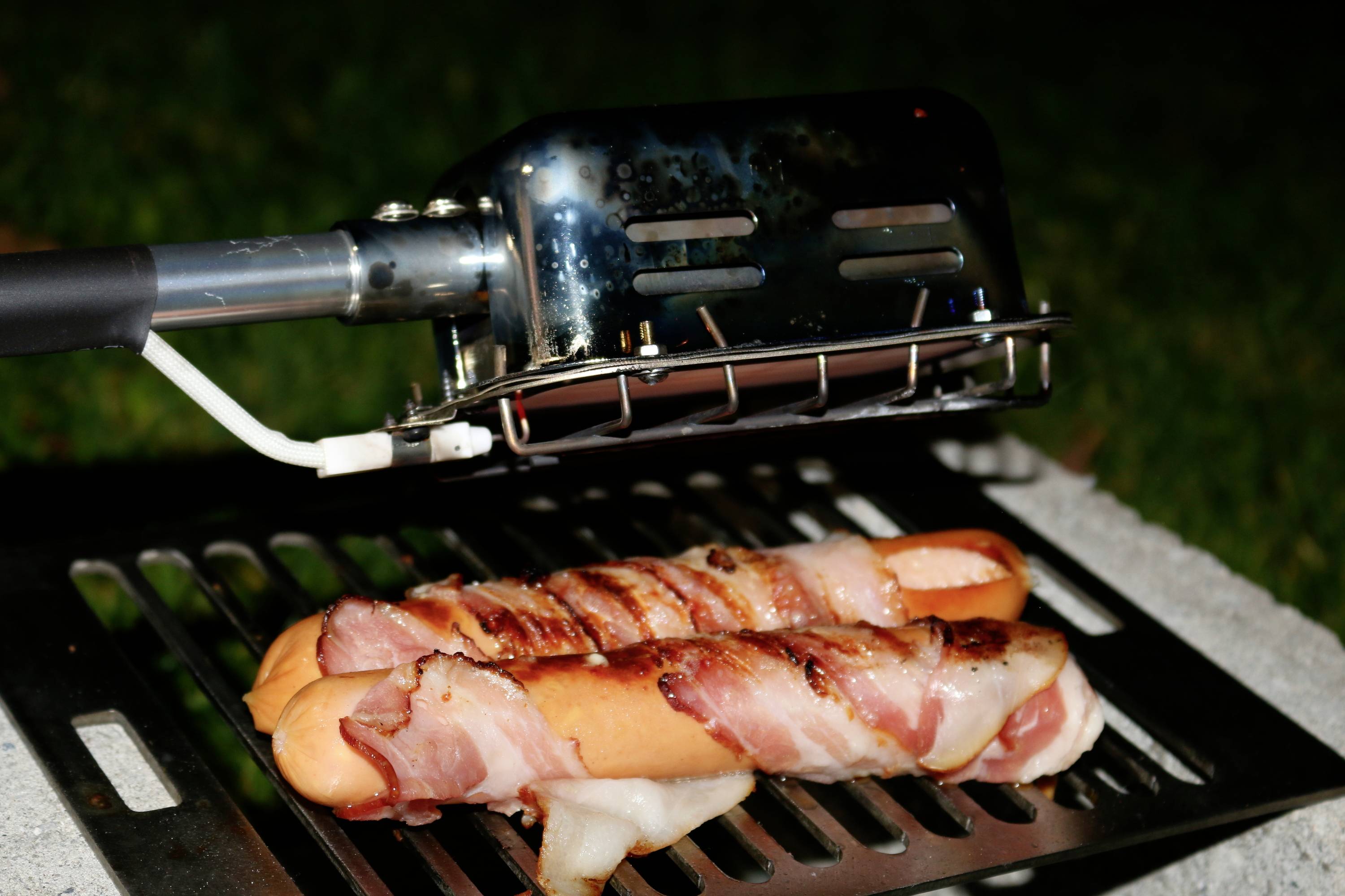 Sausages, wrapped in bacon, are grilled on a barbecue. A metal basket is used for easy handling.