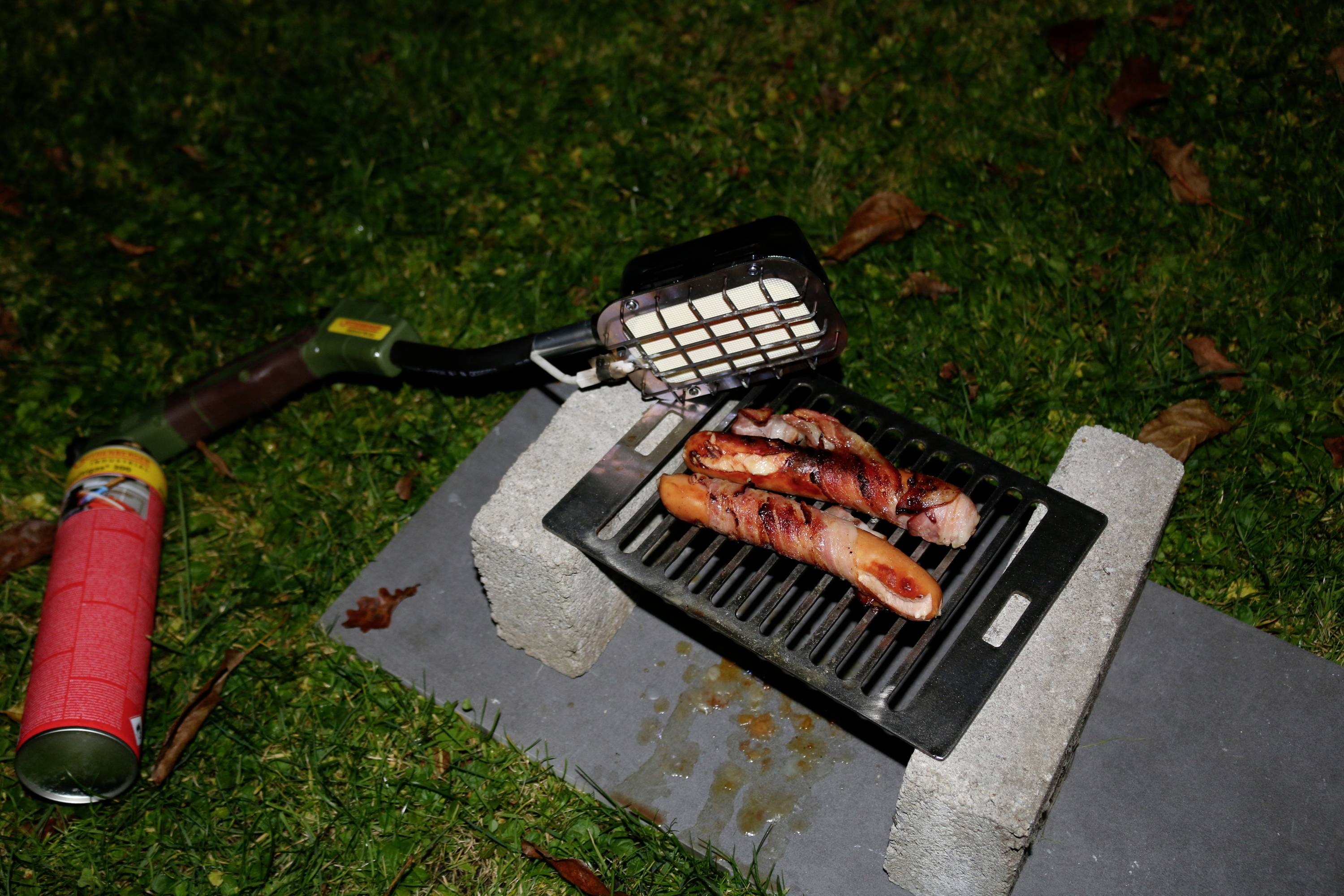 A small barbecue station on the grass. Sausages are cooking over an open flame, with a lighter tin beside them.