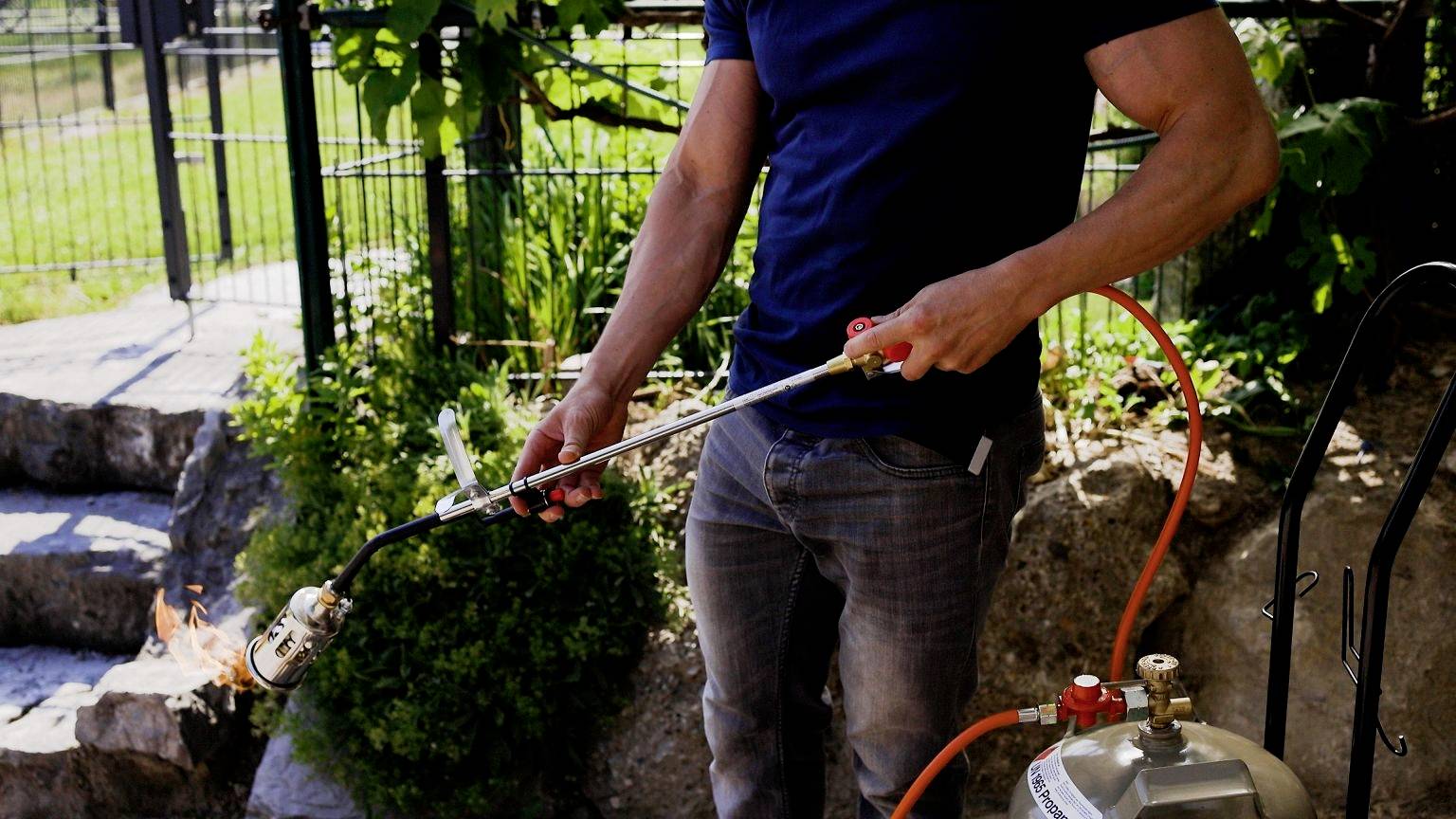 A person outdoors is holding a weed burner, connected to a gas canister, and combating weeds on a garden path.