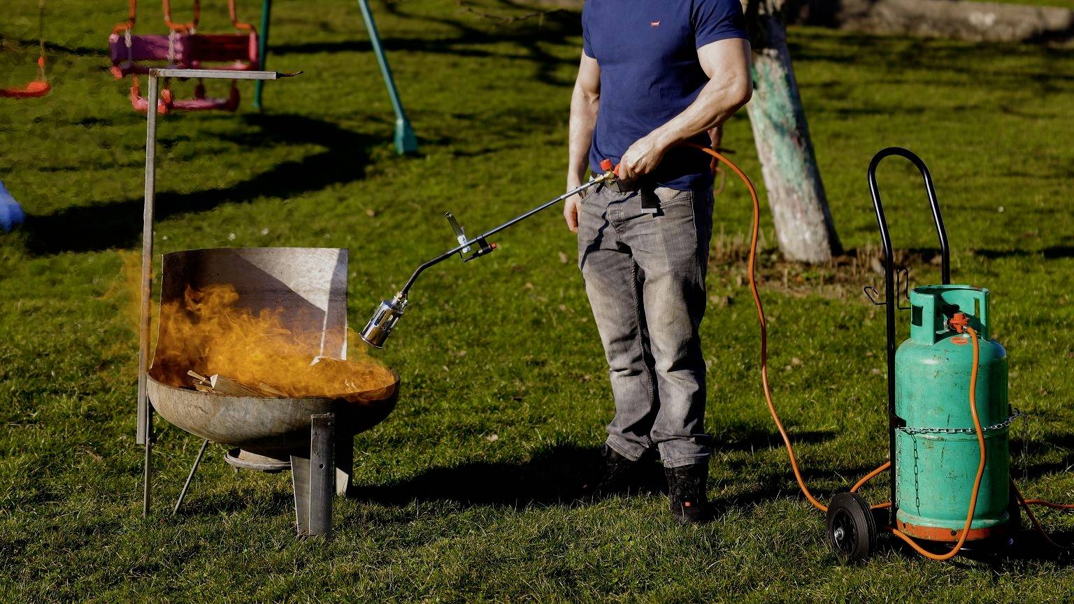 A person is standing in a garden and lighting a barbecue with a gas burner. Beside them, a gas cylinder is placed on a trolley.