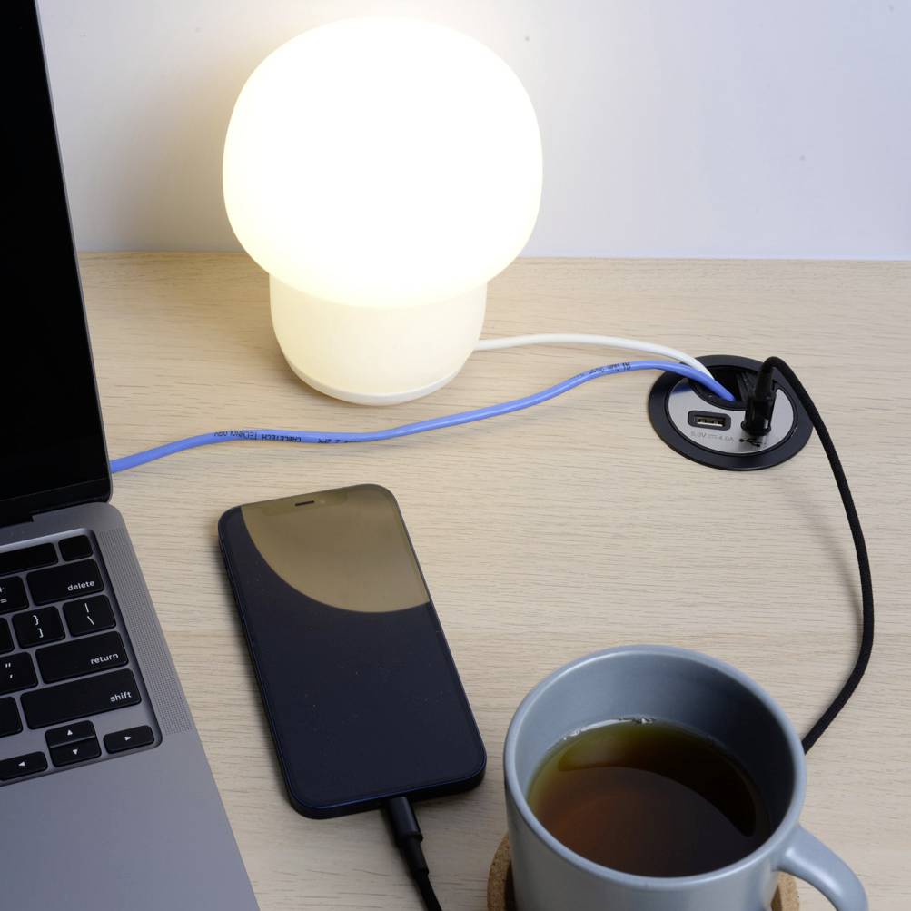 A wooden table with a glowing lamp, a smartphone, a laptop partially in view, a cup of tea, and a cable management system.