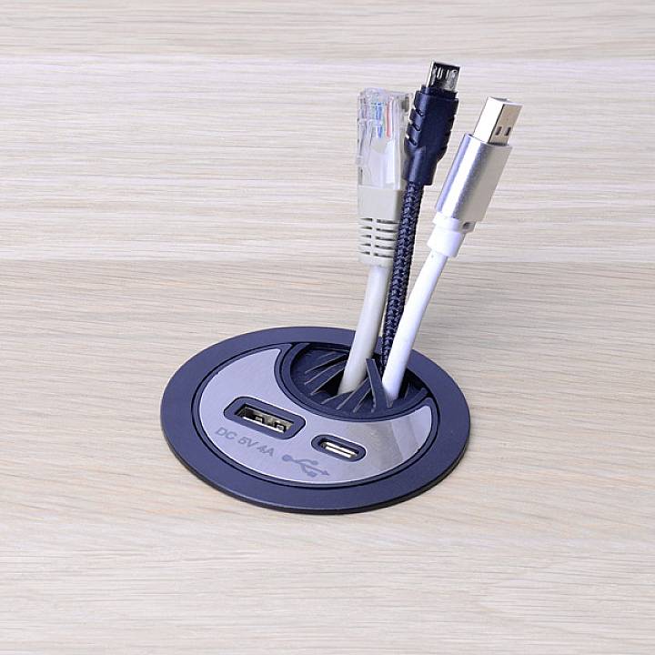 Several cables and connectors protrude from a round desk socket: USB, Ethernet and power cables on a wooden table.