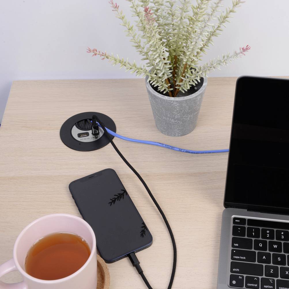 A laptop and smartphone are on a desk. Both devices are connected to charging cables. A cup of tea and a plant are sitting beside them.