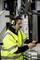 A man in a yellow high-visibility vest and safety glasses, working on a device in a factory environment, wearing headphones with a microphone.