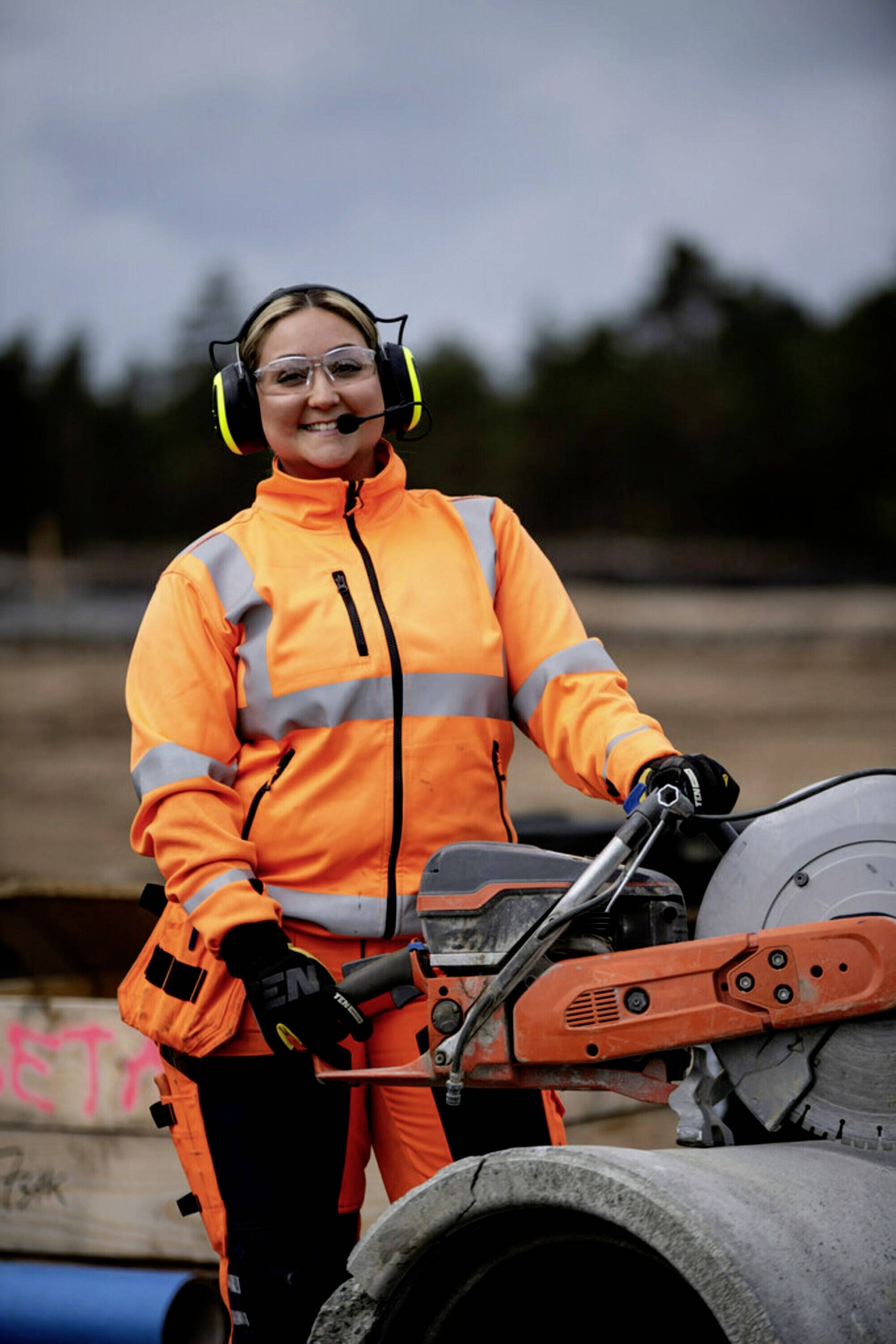 A female worker in an orange safety suit is using a circular saw to cut pipes on a construction site.